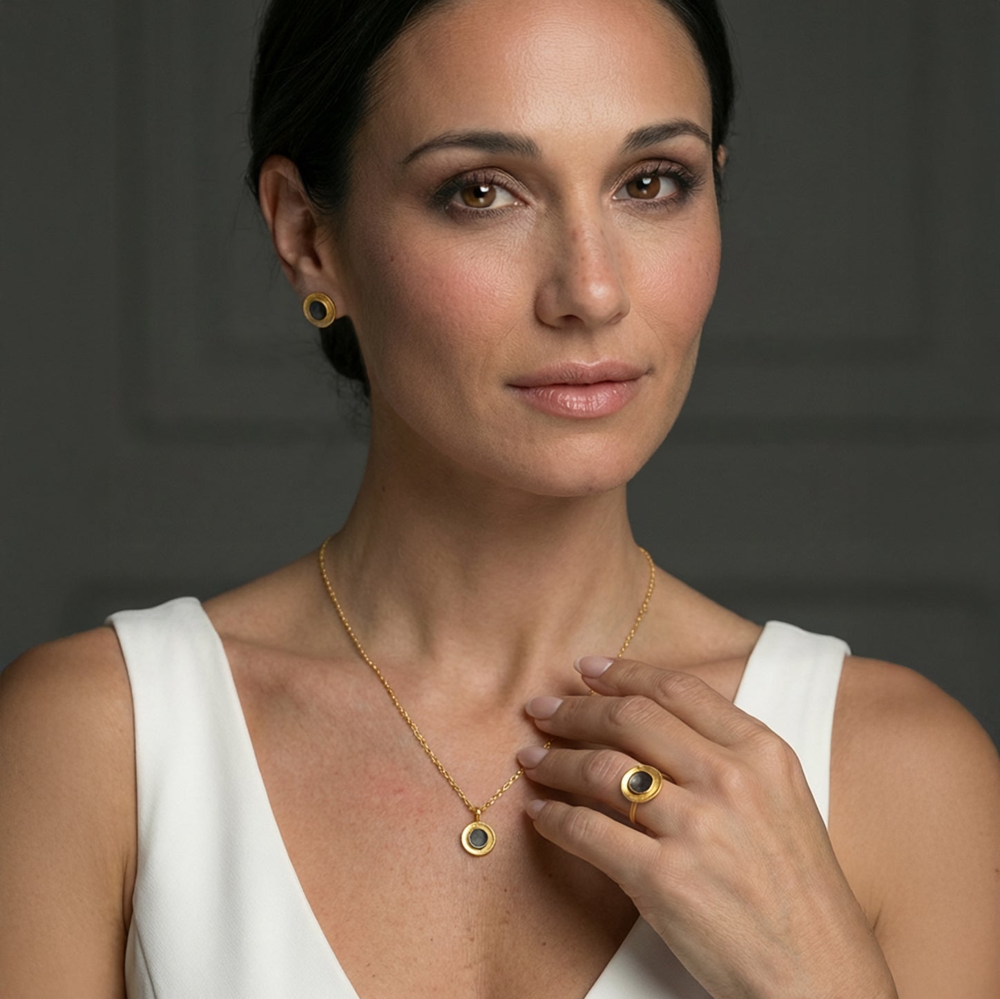 A woman wearing a gold stud earring, necklace and ring, featuring a dark patina center, photographed against a soft neutral background.