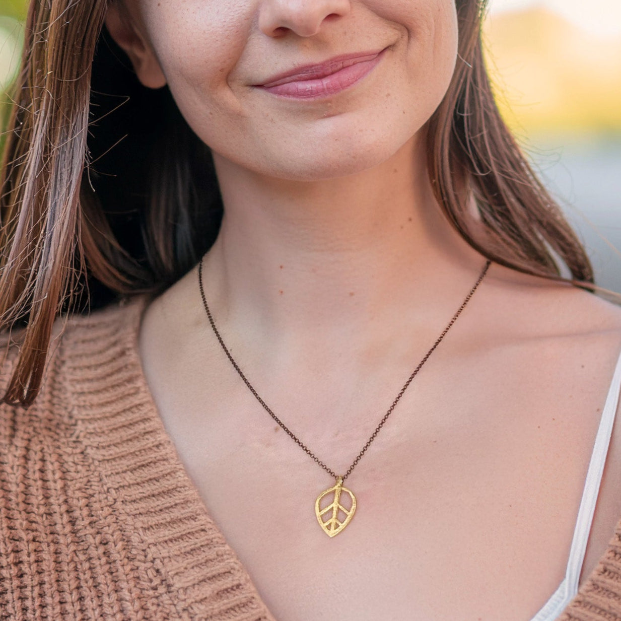 Woman wearing a brown sweater and a gold leaf-shaped pendant with a blurred outdoor background