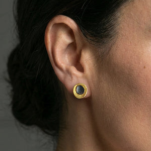 Close-up of a woman's ear wearing a small gold stud earring featuring a dark patina center, photographed against a soft neutral background.
