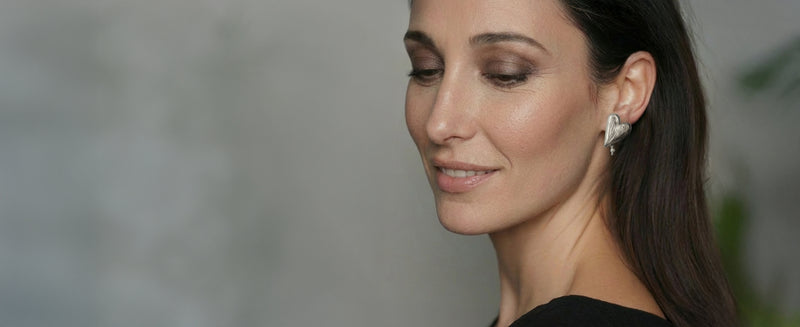 Woman wearing a heart-shaped earring with a dangling pearl, against a neutral background
